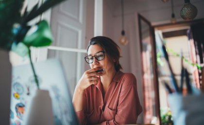 A woman in glasses sits at a desk in a home office looking worried.